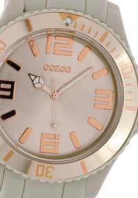 Montre à quartz avec un cadran rond argenté, des accents en or rose et un bracelet en silicone gris clair. Comprend des index horaires audacieux et une lunette en métal.