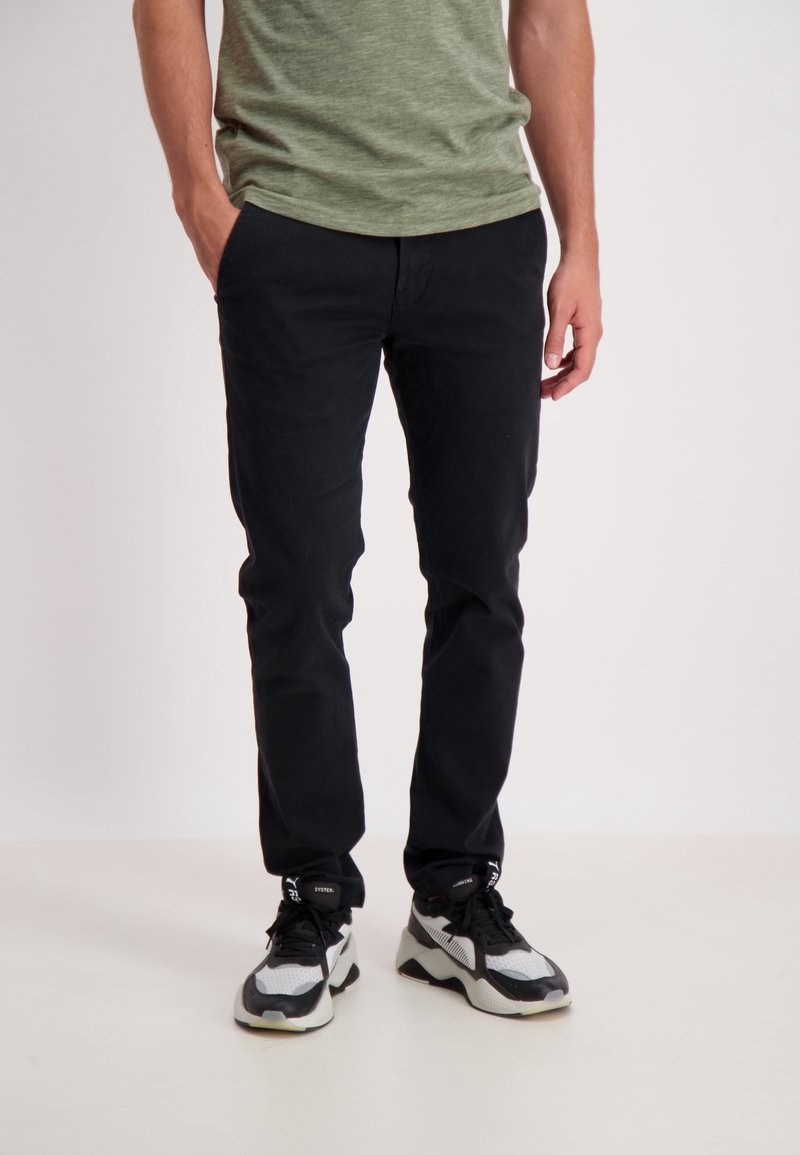 Czarne bawełniane spodnie o prostym kroju, zestawione z szaro-czarnymi trampkami. Model nosi zieloną koszulę z krótkim rękawem.