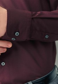 Burgundy skjorte med tekstureret finish, udstyret med blå knapper på mansjetterne, skræddersyet pasform og oprevne ærmer.