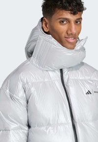 Ασημί μπουφάν τύπου puffer με ψηλό γιακά και μαύρο φερμουάρ. Κατασκευασμένο από γυαλιστερό, καπιτονέ υλικό, με το λογότυπο της Adidas στο στήθος.