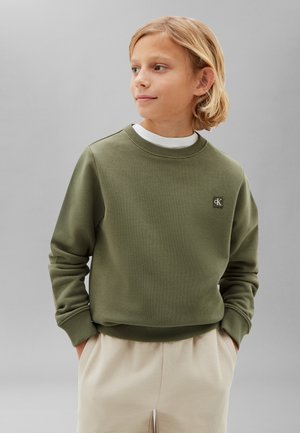 Dziecko o blond włosach ubrane w oliwkową bluzę Calvin Klein i beżowe spodnie, ręce w kieszeniach, patrzy na bok.