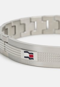 Bracelet en acier inoxydable argenté avec un bandeau texturé, présentant un petit motif triangulaire et un logo rectangulaire avec des couleurs bleu, blanc et rouge.