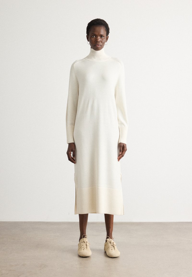Robe tricotée longue de couleur crème avec un col montant, des détails côtelés, des fentes latérales et une coupe décontractée, complétée par des chaussures beige clair.