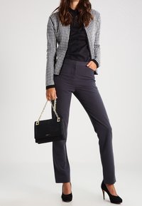 Svart och vit houndstooth-blazer, svart blus, grå anpassade byxor, svarta mockaskor med klack, samt en kviltad svart handväska med kedjestropp.