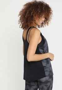 Kvinna med lockigt hår som bär en svart och grå t-shirt utan ärmar med batikmönster och matchande leggings, leende och vänd lite på sidan.