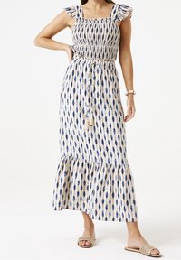 Sukienka maxi z niebieskim wzorem liści na beżowej tkaninie, gorsetem na gumce, falbaniaste ramiączka i warstwową spódnicą. Detal z frędzlami w talii.