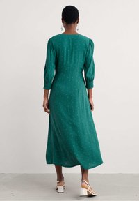 Vestido verde de manga larga con cintura ajustada, longitud a media pantorrilla y patrón de lunares blancos. Tela suave con un ligero brillo.