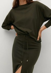 Kvinde iført en olivengrønn langærmet top, der er stukket ned i en matchende høj taljeret nederdel med frontslids og snøre i taljen.