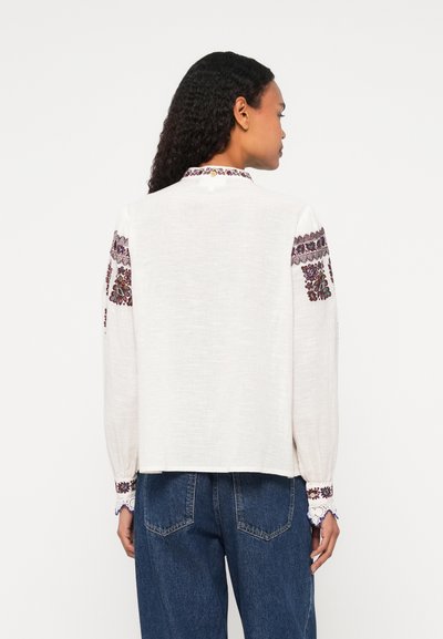 Fehér vászonblúz, hímzett díszítéssel az ujjakon és a galléron, lazán illeszkedő fazonnal és hosszú, lebegő ujjakkal.