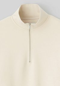 Sweatshirt zippé beige clair avec un col haut, texture douce et poignets côtelés. Doté d'une fermeture éclair métallique au centre devant.