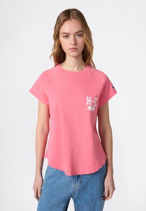 Junge Frau mit langen blonden Haaren, die ein rosa kurzärmliges Shirt mit der Aufschrift "N-E 7" auf der Brust und blaue Jeans trägt, vor weißem Hintergrund.