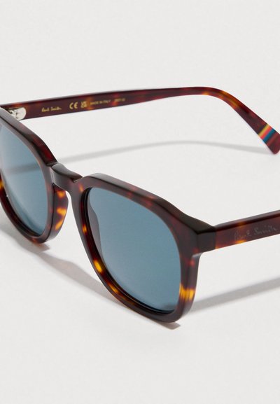 Ochelari de soare cu rame tip țestoasă, cu lentile albastre, un cadru gros și un design geometric. Prezintă un logo metalic „Paul Smith” pe braț.