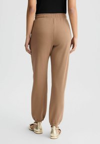 Braune, zulaufende Sweatpants mit elastischem Bund und Bündchen. Glatte Stofftextur. Getragen mit hellgoldenen Sandalen.