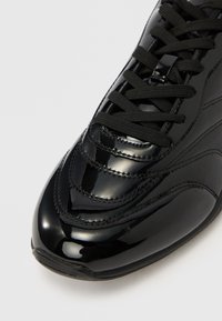 Μαύρο αθλητικό παπούτσι από δέρμα πατέντα, με υφ ή ραφών, επίπεδα μαύρα κορδόνια, στρογγυλερή μύτη και λείο φινίρισμα. Μινιμαλιστικός σχεδιασμός, χωρίς ορατά λογότυπα.