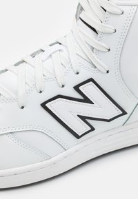 Bílé sportovní boty s texturovaným koženým svrškem, který má černé akcenty a výrazné bílé logo "N". Šněrování je propletené skrze očka.