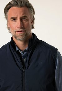 Mörkblå, ärmlös väst i slät tyg med dragkedjestängning. Bärs över en ljusblå skjorta med knappar och har subtil logotypdetalj.