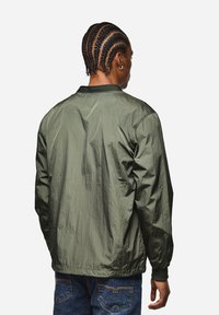 Olivegrüne Bomberjacke mit einer leicht zerknitterten Textur, geripptem Kragen und Bündchen. Sie hat eine lockere Passform und lange Ärmel.