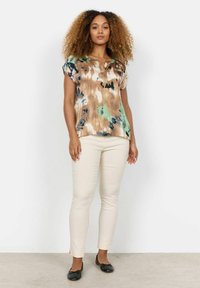 Kurzarmige Bluse in Braun, Grün und Schwarz mit Batikmuster und ausgeschnittenem Ausschnitt, kombiniert mit cremefarbenen Slim-Fit-Hosen und schwarzen Ballerinas.
