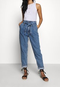 Vaaleansiniset korkeavyötäröiset farkut, joissa on sidottava vyötärö ja käännetyt lahkeet, yhdistettynä valkoiseen hihattomaan puseroon ja mustiin remmisandaaleihin.