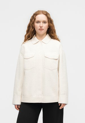 Pitkä, kihara punapäinen nainen, jolla on teksturoitu, valkoinen takki, jossa on läppätaskut rintakehässä, ja mustat housut, seisomassa yksinkertaisen taustan edessä.