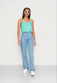 Top verde claro, sem mangas, com botões frontais e detalhe de laço, acompanhado de jeans claros de cintura alta e ténis brancos. Óculos de sol brancos incluídos.