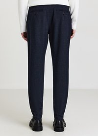 Marineblaue, strukturierte Hose mit zwei Gesäßtaschen und schwarzen Knöpfen. Eng zulaufendes Bein mit Bündchen an den Knöcheln, kombiniert mit einem weißen Pullover.