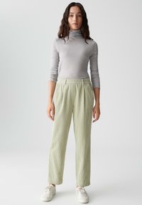 Maglione a collo alto grigio chiaro e pantaloni a gamba larga di un verde chiaro con texture, dotati di vita elasticizzata. La modella si trova su uno sfondo neutro.