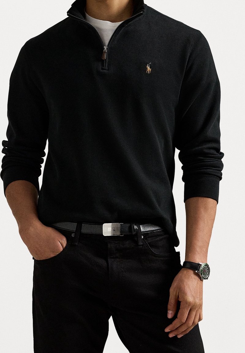 Homme portant un pull noir à fermeture éclair quart avec un petit logo, un sous-vêtement blanc, un pantalon noir, une ceinture noire et une montre noire au poignet gauche.