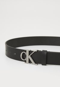 Čierny kožený opasok so striebornou kovovou sponou s logom "ck" a nastaviteľnými otvormi na veľkosť. Hladká textúra a klasický dizajn.