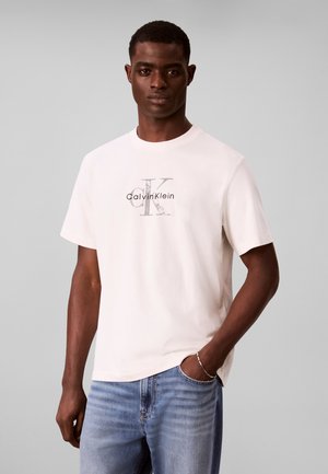 Άνδρας που φοράει λευκό t-shirt Calvin Klein και ανοιχτό μπλε τζιν, στέκεται με το ένα χέρι στην τσέπη του μπροστά σε απλό γκρι φόντο.