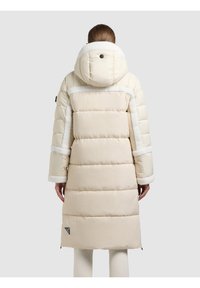 Kvinna i en lång beige vadderad jacka med huva och vit fleecekant, stående med ryggen mot en enfärgad grå bakgrund.