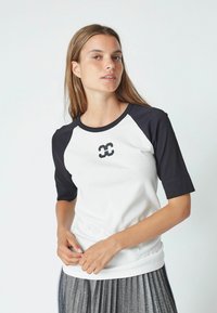 Balta ir tamsiai mėlyna raglan stiliaus marškinėliai su trumpomis rankovėmis, su unikaliu juodu logotipu ant krūtinės. Minkšta medžiaga ir prigludusi forma.