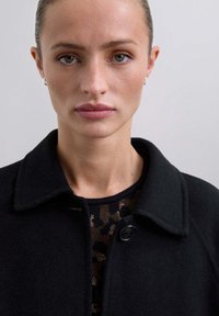 Abrigo de lana negro con cuello de solapa, que presenta un cierre de un solo botón. Debajo, se ve un suéter oscuro con estampado.