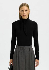 Zwarte langemouwtop met een hoge nek en strikdetail. Gecombineerd met hooggepolsterde grijze gestreepte broeken, met een losse pasvorm en plooien.