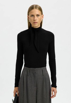 Czarna bluzka z długim rękawem, wysokim kołnierzem i detalem w postaci wiązania. Połączona z szarymi spodniami o wysokim stanie, w wzór, charakteryzującymi się luźnym krojem i plisami.