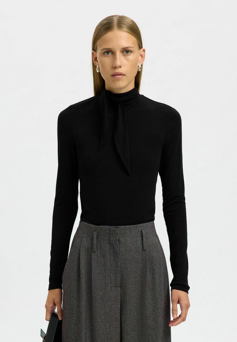 Zwarte langemouwtop met een hoge nek en strikdetail. Gecombineerd met hooggepolsterde grijze gestreepte broeken, met een losse pasvorm en plooien.