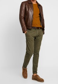 Veste en cuir marron portée sur un pull en maille texturée couleur moutarde, associée à un pantalon chino vert olive et des chaussures en daim marron, créant un look superposé.