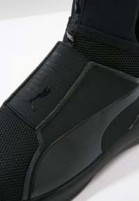 Musta urheilukenkä, jossa on verkkokangas, joustava hihna Puma-logolla ja tyylikäs synteettinen sivupaneeli valkoista taustaa vasten.