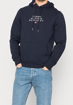 Hombre que lleva una sudadera con capucha Tommy Hilfiger azul marino con texto y logo blancos, combinada con vaqueros azul claro, manos en los bolsillos.