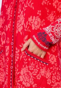 Roter Strickcardigan mit Blumenmustern, rosa Knöpfen, Taschen und kontrastierenden weiß-blauen gemusterten Bündchen.