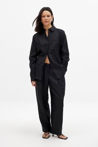 Črna lanena srajca in hlače širokega kroja, ki vključujejo gumbasto zapenjanje spredaj, eno žep na prsih in sproščen kroj. Gladka tekstura.