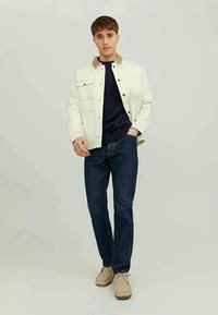 Casaco de ganga branco com colarinho bege sobre um suéter azul marinho, combinado com jeans azul escuro e sapatos bege. Design simples, ajuste descontraído.
