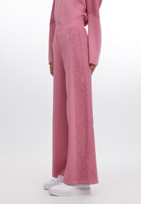 Rosa vide benklær med blomsterlacemotiver på sidene, laget av et glatt stoff. Akkompagnert av hvite joggesko.