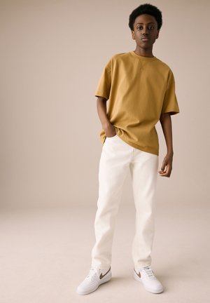 Jaunas žmogus stovi kišenėje ranka, dėvi garstyčių spalvos per dydį didelį marškinėlį, baltas kelnes ir baltus sportbačius su rudu logotipu.