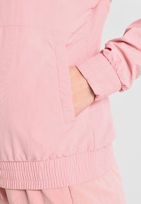 Light pink windbreaker featuring a front pocket, elastic cuffs, and a gathered waistband, made from a smooth, lightweight fabric.