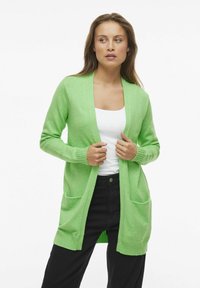 Grön cardigan med avslappnad passform, öppen front, ribbade ärmslut och två framfickor. Bärs över en vit topp och tillsammans med svarta byxor.