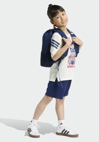 Tmavomodrý batoh s ramennými popruhmi, nosený cez krémovú košeľu so Spider-Man grafikou, doplnený tmavomodrými šortkami a bielymi teniskami.