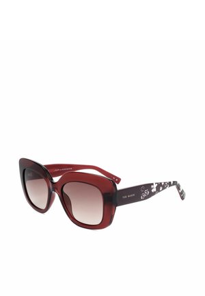 Occhiali da sole oversize a forma di occhio di gatto rossi e neri con lenti sfumate e motivo floreale sulle aste, marchiati Ted Baker sulla stanghetta.