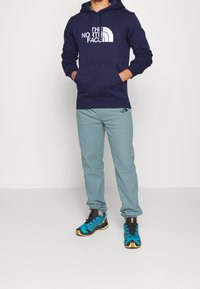 Felpa della marina con logo bianco, pantaloni della tuta blu chiaro e scarpe da atletica nere/blu con design angolare; marchio chiave ben visibile sugli indumenti.