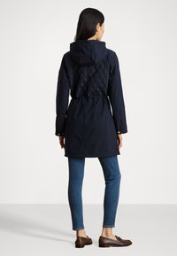 Parka navy con retro imbottito, vita regolabile e cappuccio. Presenta maniche lunghe e dettagli in bottone dorato, abbinata a jeans blu e scarpe marroni.